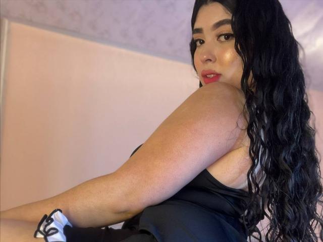 AgathaLynx with long black wavy hair, red lipstick, and black tank top, showing her back and bare shoulder, sitting with her ass slightly raised. Wearing a white scrunchie.