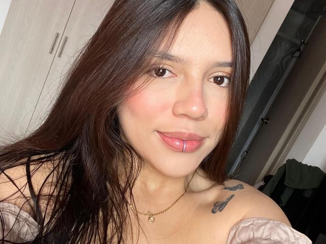 SophiaMarx has long brown hair, pink lips with a labret piercing, light skin, and small tattoos on her right shoulder. She's wearing a pink off-shoulder top.