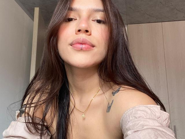 SophiaMarx with long brown hair, pink off-shoulder top, light skin, small gold necklace, subtle tattoos on shoulder, pouty pink lips, slightly squinted eyes, soft natural light.