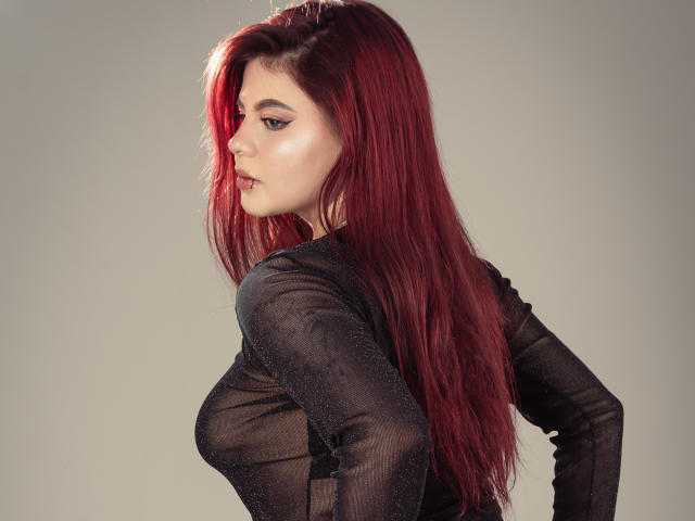 MelanieGate with red hair, black sheer top, side profile, showing her tits and ass, looking serious, light skin, makeup, and long hair against a neutral background.