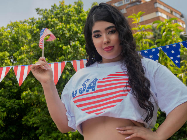 AgathaLynx with curly black hair, holding a small American flag, wearing a white crop top with red and white stripes, exposing her large tits, standing in front of green trees.