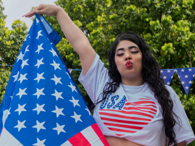 AgathaLynx with long black hair, puffy red lips, white USA shirt, heart pattern, holding American flag, trees in background, slightly overweight, medium tits, soft light, daytime.