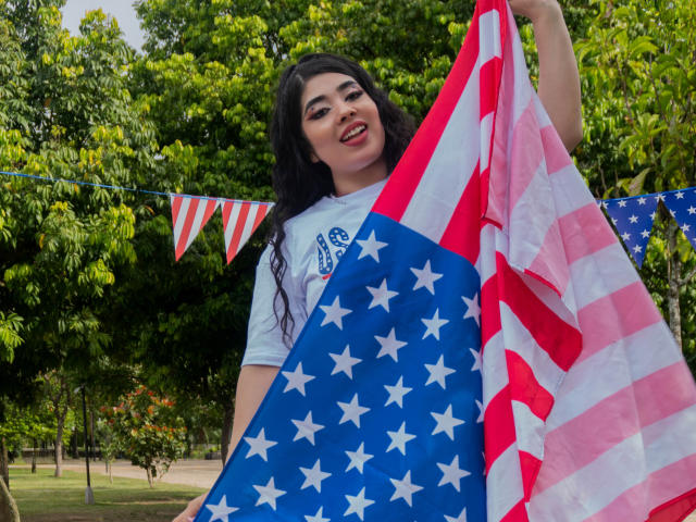 AgathaLynx with long black hair, smiling, holding two American flags. She's wearing a white shirt, standing outside, with red-white-blue bunting in the background. Her tits are covered.