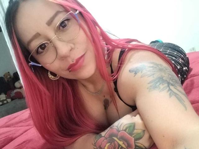 Lauren Blake, con i capelli rosa, gli occhiali, il piercing al naso e al labbro, sdraiata su un letto rosa, mostra tatuaggi sul braccio e sul petto, indossa un vestito nero e guarda la fotocamera..