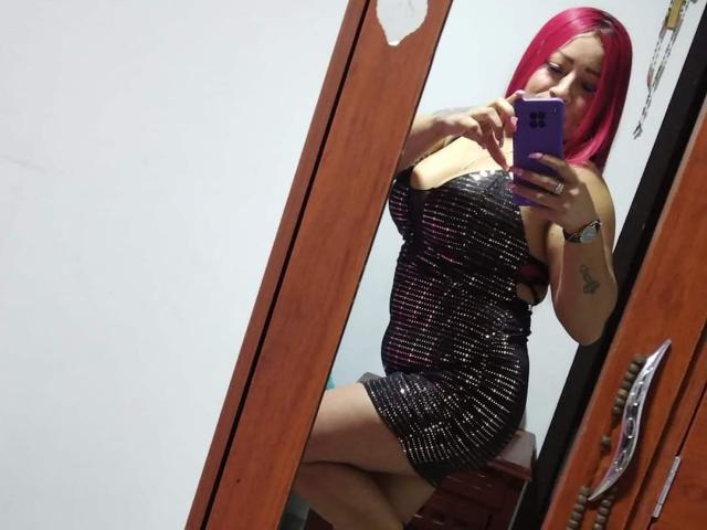 Lauren Blake, met roze haar, draagt een glinsterende zwarte jurk en zit voor een spiegel. Ze houdt een paarse telefoon vast en toont haar borsten en billen. Ze heeft een tatoeage op haar linkerarm..