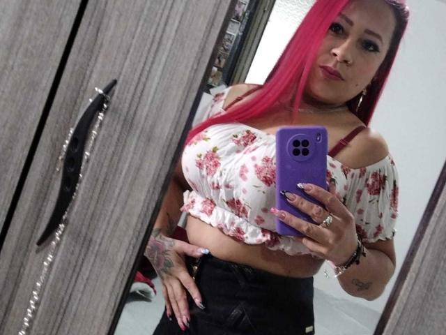 LaurenBlake with pink hair, off-shoulder floral top, showing her boobs, holding a purple phone, black pants, tattoos, ring, mirror selfie.