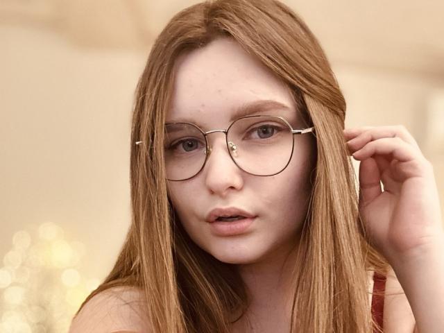 MagikPeachh has long brown hair, wearing round glasses with thin metal frames, looking slightly surprised with her hand near her face, showing her cute, pale face and small nose.