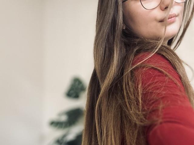 MagikPeachh con cabello largo y castaño, camisa roja, gafas y una sonrisa sutil, mostrando un perfil lateral. Los senos no son visibles, el enfoque está en el rostro y el cabello. Luz suave y natural..