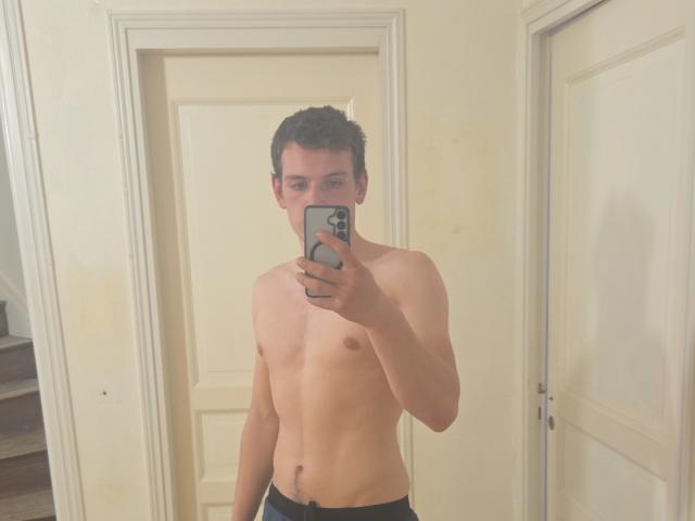 MarioDiamond, shirtless, holding a phone in front of a mirror, shows a slim, toned, light-skinned body with a small, unerect, uncircumcised penis, wearing black pants.