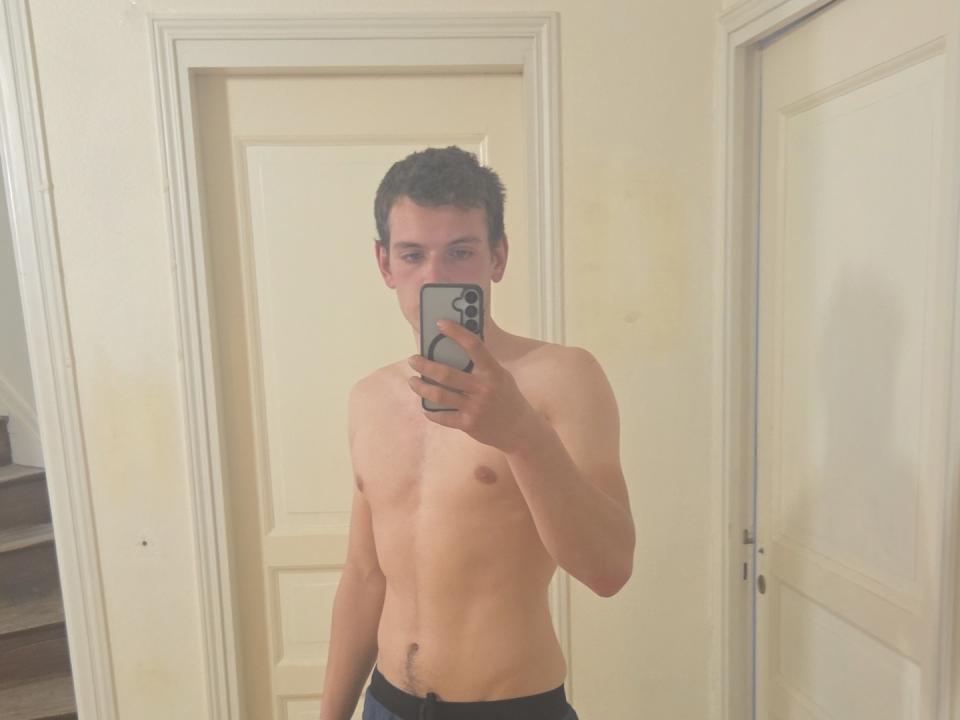 MarioDiamond, shirtless, holding a phone in front of a mirror, shows a slim, toned, light-skinned body with a small, unerect, uncircumcised penis, wearing black pants.