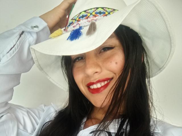 Madisson Fontaine, avec ses cheveux noirs, son rouge à lèvres rouge, son chapeau blanc orné de plumes colorées et sa chemise blanche, sourit à la caméra, une main tenant le chapeau, fond blanc, tête légèrement inclinée..