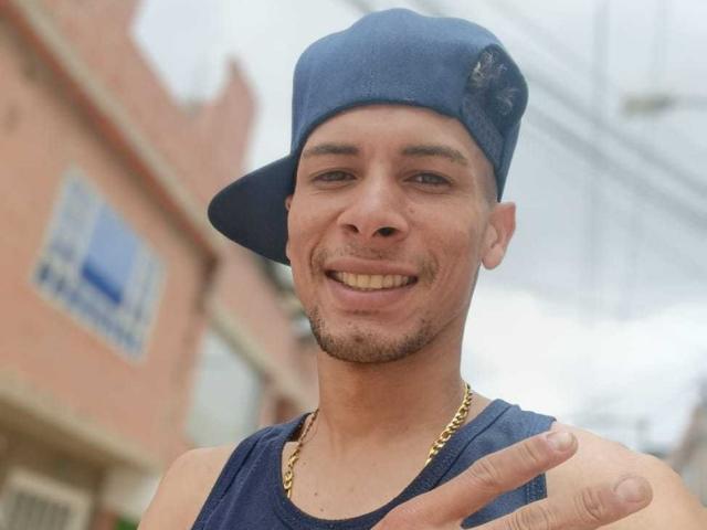 Fornicatorass with medium skin, blue cap, gold chain, and dark tank top, smiling, pointing with right hand, short beard, and visible teeth. Overcast sky in background.