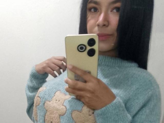 Fornicatorass with dark hair, holding a yellow iPhone, wearing a light blue sweater with beige floral patterns, taking a selfie. Boobs visible under sweater.
