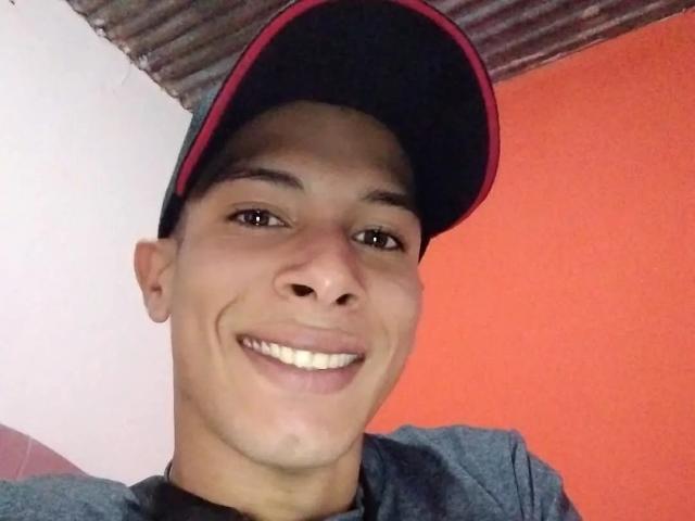 Fornicatorass with a cute smile, wearing a black and red cap, grey shirt, and dark shirt underneath. Short, dark hair, brown eyes, and a slightly tanned complexion.