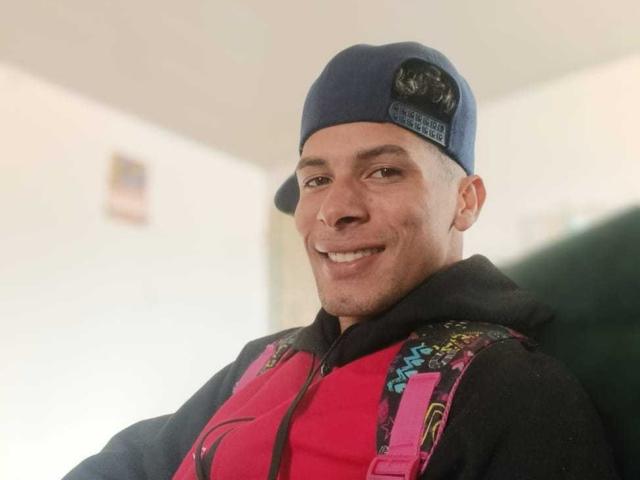 Fornicatorass wearing a blue backward cap with a black logo, smiling, dressed in a pink and black hoodie with colorful straps. Light-skinned, short-haired, slightly stubbled.