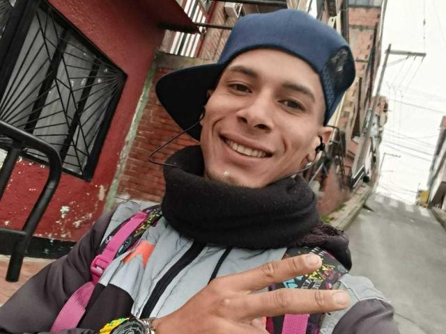 Fornicatorass with a blue cap, black scarf, colorful jacket, and earbuds, smiling, showing a finger to the camera, has a cute, thin face, and is wearing pink straps.