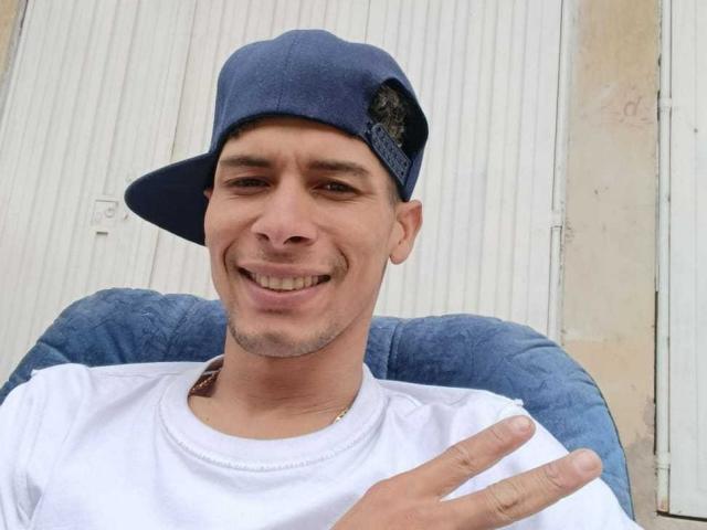 Fornicatorass, smiling with brown eyes, short dark hair, wearing a blue cap and white tee, making a peace sign with right hand.