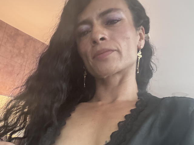 NataliaKnox with long black hair, purple eyeshadow, small nose ring, gold earrings, black lace top, slight smile, looking down, light skin, slight wrinkles, soft lighting.