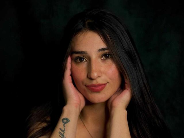 ElecttraKane has dark, straight hair, brown eyes, and a tattoo on her right wrist. She's touching her face with both hands, smiling softly. Dark green background.