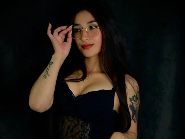 ElecttraKane with long black hair, glasses, red lipstick, black lace dress, tattoos on arm and wrist, holding glasses, looking at camera, dark background. Boobs visible, soft smile.
