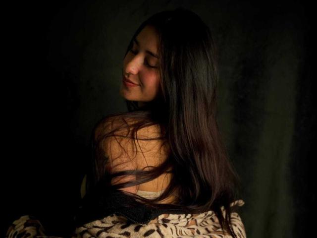 ElecttraKane with long dark brown hair, eyes closed, soft smile, wearing a leopard-print top, black straps falling off shoulders, dark greenish-black background, gentle lighting.