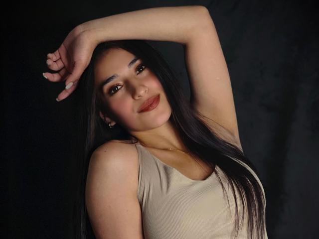 ElecttraKane with long black hair, tan sleeveless top, raised arm, slight smile, brown eyes, red lipstick, cute, tan skin, small gold necklace, dark background, sexy, attractive.