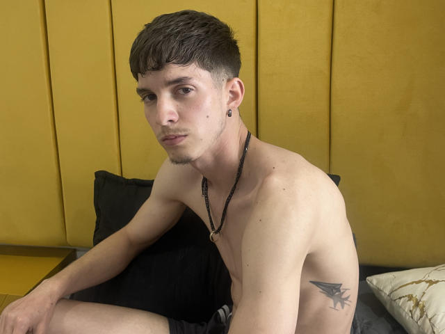 Shirtless JheamPiero with short brown hair, black ear plug, necklace, small tattoo on right side, sitting on black pillow, yellow wall in background, looking at camera, slightly pouty.