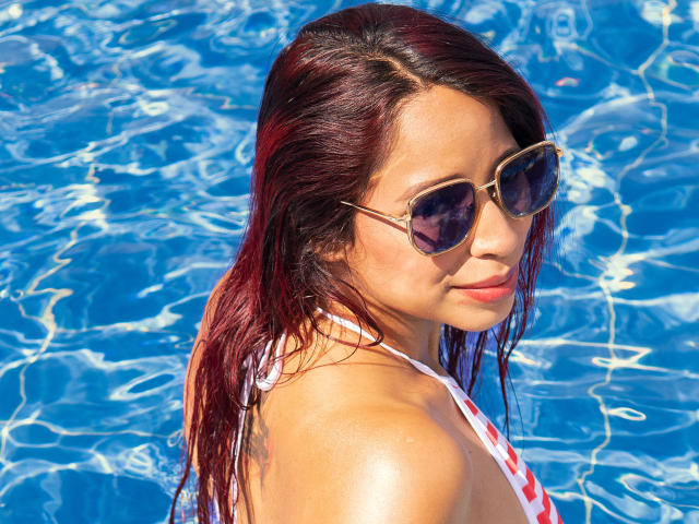 VivianCruzz with wet red hair, wearing sunglasses, and a white-red bikini top, standing in a blue pool, looking back with a slight smile. Her tits are visible.