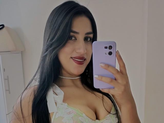 AlanaHarris with long black hair, wearing a lace bra showing her tits, pearl necklace, and red lipstick, taking a selfie with a purple phone, smiling.