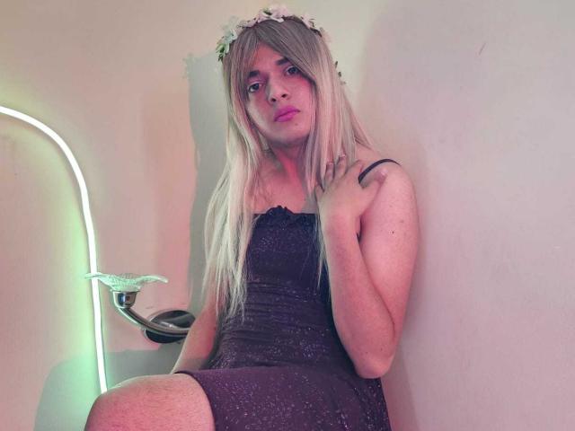 GeciaLove with long blonde hair, wearing a sparkly black dress, flower crown, and pink makeup, sitting with one hand on her chest, neon light in background.