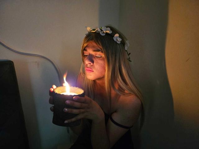 GeciaLove with long blonde hair, flower crown, and black strap dress, holding candle, eyes closed, soft candlelight, gentle shadow on face, dimly lit, serious expression, warm glow.