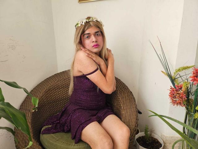 GeciaLove, a blonde girl with a flower crown, sits in a wicker chair, wearing a purple dress, with arms crossed, looking surprised, and has pink lipstick.