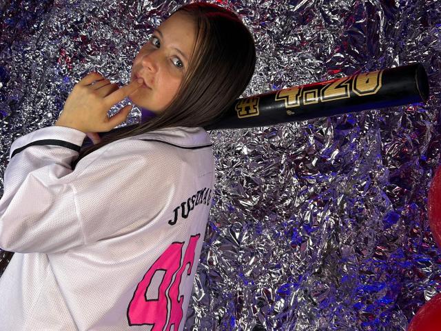 GraceRedMiller with brown hair, white baseball jersey, holding black and gold bat, biting finger, looking at camera, silver foil background, slightly smiling.