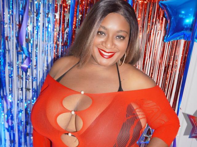 Nahomi Anders, con un seno prosperoso, indossa una maglia rossa in rete con aperture, sorride alla fotocamera, con rossetto rosso, capelli castani, orecchini a cerchio, in piedi di fronte a decorazioni blu e rosse..