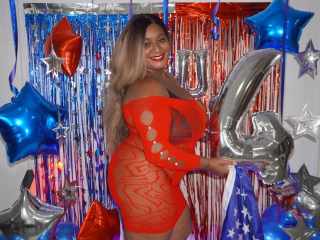 NahomiAnders, with big ass and red see-through dress, holding a silver "2" balloon, smiling, standing in front of colorful stars and metallic strips, has long blonde hair.
