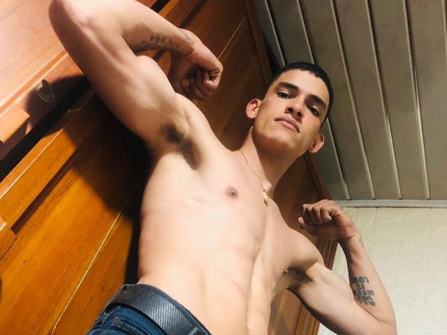 CarlosJaramillo, shirtless with defined muscles, flexes arms, showing armpit hair, and has tattoos on right forearm, wearing jeans, looking down, in a wooden room with metal ceiling.