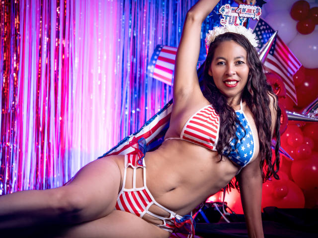 RosalindaCortez in a red, white, and blue bikini with American flag patterns, lying down with arm raised, smiling, wearing a white headband, showing her tits and ass.