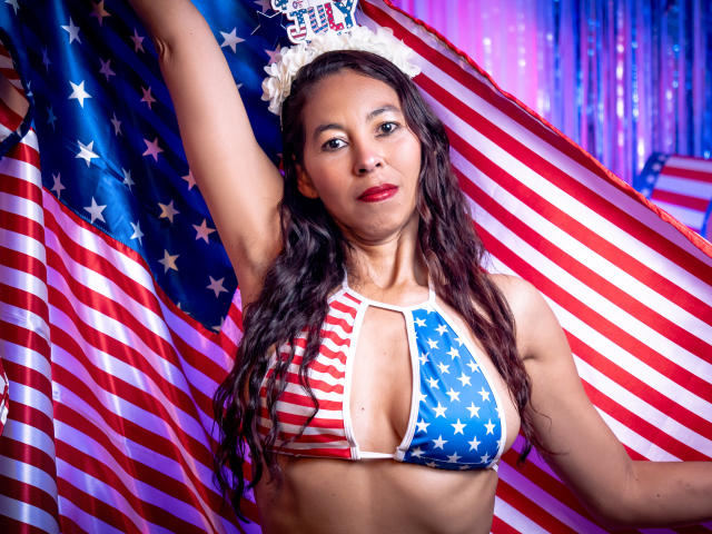 Rosalinda Cortez in einem bikiniartigen Oberteil mit amerikanischen Sternen und Streifen, die eine riesige amerikanische Flagge hält, mit langen, welligen Haaren, rotem Lippenstift und einer weißen Krone, wobei ihre Brüste zu sehen sind und sie ein selbstbewusstes Lächeln zeigt..