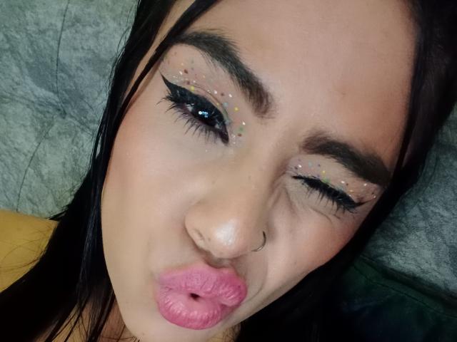 StellaBeckker with pink pout, black eyebrows, glittery eyeliner, and nose ring, puckering her lips, has dark hair, and a cute, sexy face, looking up with half-closed eyes.