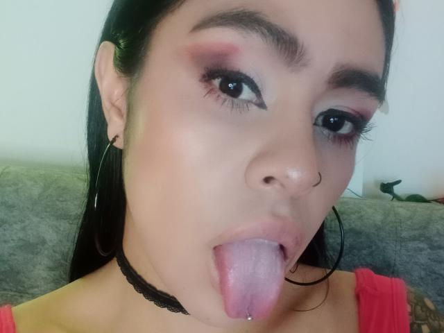 StellaBeckker's face with black hair, hoop earrings, nose ring, pink eyeshadow, black choker, tongue out, showing tongue piercing, and a small tattoo on her shoulder.