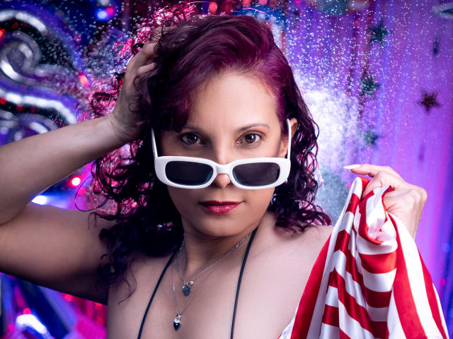 AdaMclain with curly purple hair, white sunglasses, and a red-and-white striped top, holding it with one hand, showing off her tits and necklace with a purple gem.