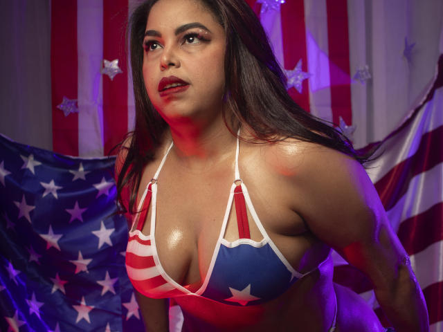 MorganMoretz with shiny skin, wearing a star-spangled bikini that exposes her big, sweaty tits, looking up with red lipstick and dark brown hair, against American flags.