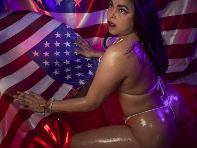 MorganMoretz with shiny, oiled skin, wearing a tiny white thong, holding a large American flag, purple lighting, looking back with red lipstick, and bracelets on her right wrist.