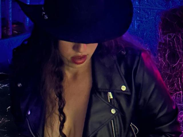 RachelWildest with a black witch hat, dark red lipstick, and curly hair, wearing a black leather jacket, exposing her tits. Blue and purple lights create a sexy, mysterious vibe.