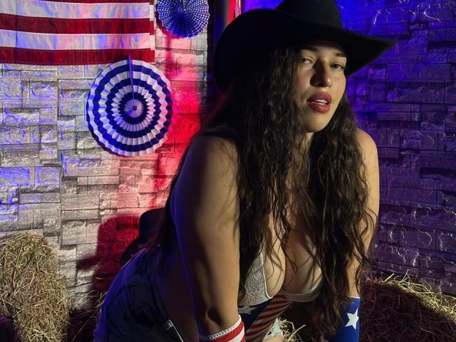 RachelWildest with long curly brown hair, wearing a black cowboy hat, white lace top, and blue jeans, leaning forward with visible large boobs, American flag decor, and blue lights.