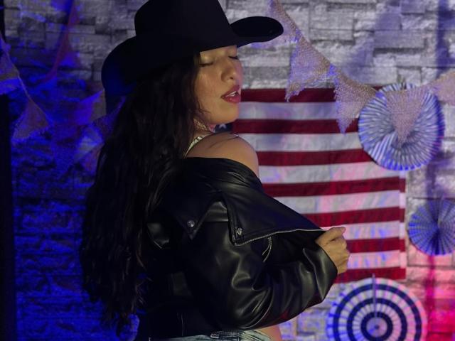 RachelWildest with long black hair, wearing a black hat and leather jacket, showing her bare shoulders and ass, eyes closed, red lipstick, against a colorful, patterned background.
