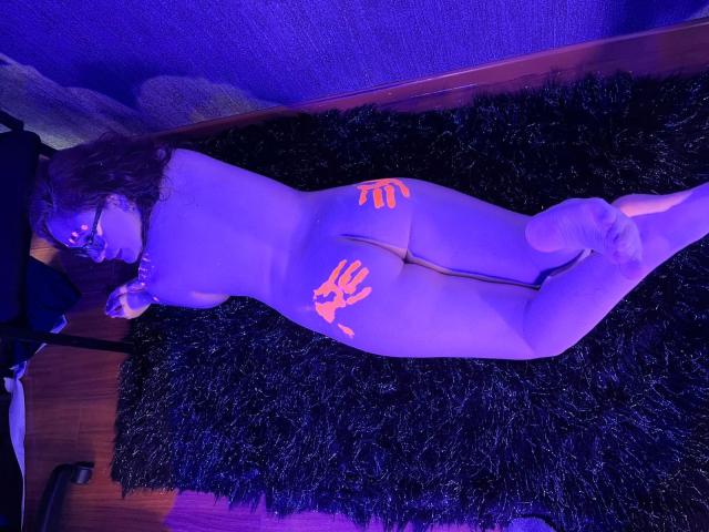 AlanisSweet, naked, lying on a black shaggy rug, illuminated by purple light, with glowing orange hand prints on her back and ass. Her curly hair and glasses are visible.