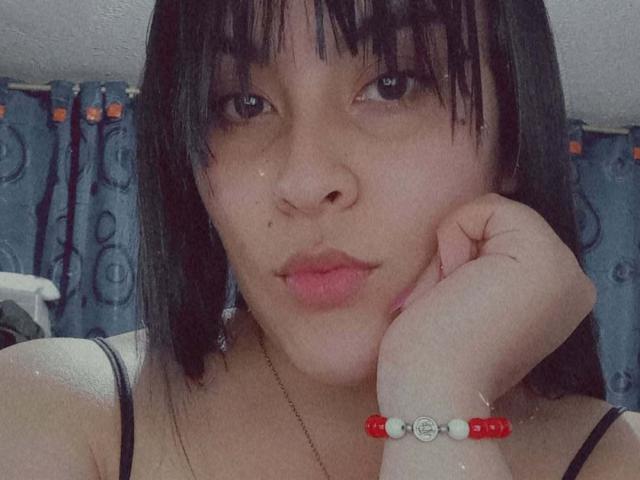 SelenaF with black hair, bangs, and dark eyes, resting her chin on her hand, wearing a red and white bead bracelet, with pink lips and a cute, small nose.