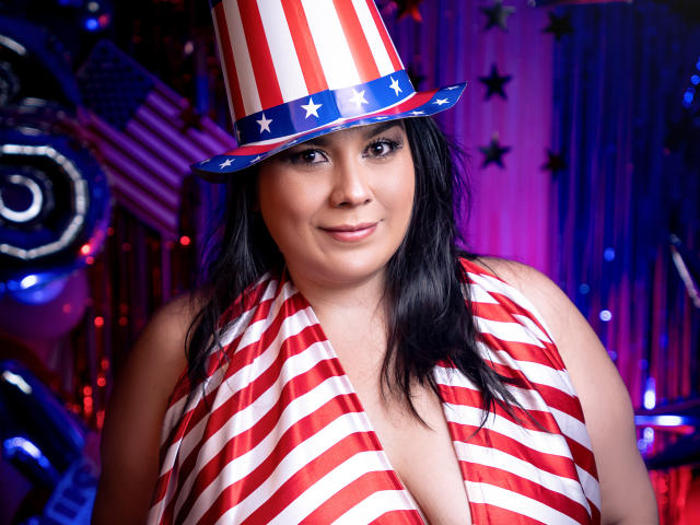 MartinaFox69, with black hair and red-white-striped top, wears a red-white-striped hat with stars, smiling, showing cleavage. Her tits are visible under the top. She's curvy.