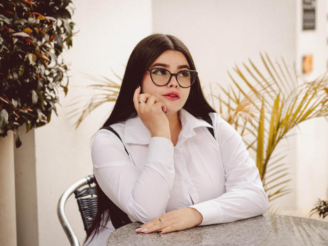 BrithneyLee with long black hair, glasses, white shirt, hand on cheek, resting on gray table, looking slightly to the side, red lipstick, small ring on right hand, pensive expression.